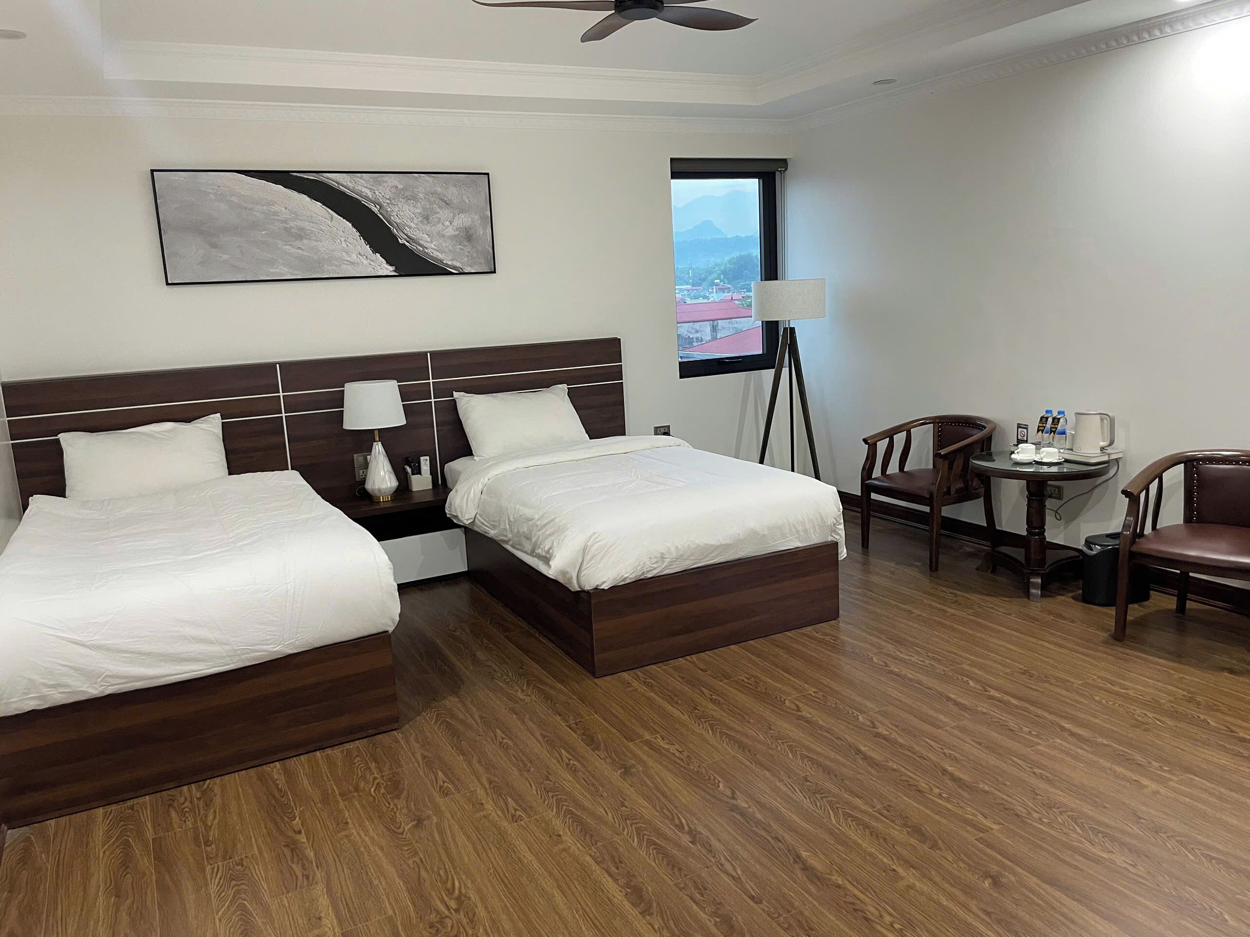 A hotel room with two beds, a desk, a chair, a lamp, and a view of a mountain through a window.