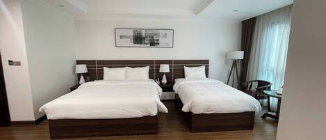 A hotel room with two beds, a ceiling fan, a bedside table with lamps, a floor lamp, and a painting on the wall.