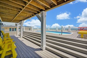 A wooden deck with steps leading to a pool area, a covered seating area, and a view of the beach.