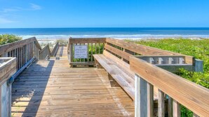 Une promenade en bois mène à la plage, et une pancarte indique : « SURVEILLANT DE BAIN DE SERVICE, BAIGNEZ À VOS PROPRES RISQUES. »