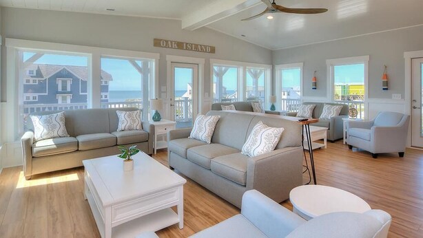 Un vaste salon doté d’une grande fenêtre offrant une vue sur les maisons et la mer, d’un ventilateur de plafond et d’une enseigne indiquant « OAK ISLAND ».