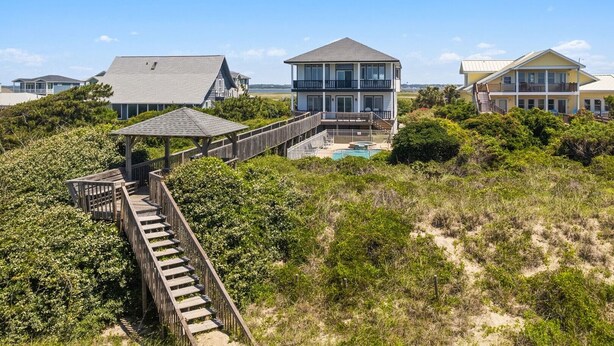 A beachfront property with a wooden deck leading to a house with a pool. - Discover Peace on the Beach, a stunning 6BR, 5BA oceanfront retreat in Oak Island. (Oak Island)