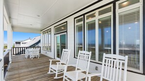 A covered outdoor deck with white rocking chairs and a table, large windows with blinds, and a view of a house in the background. - Discover Peace on the Beach, a stunning 6BR, 5BA oceanfront retreat in Oak Island. (Oak Island)