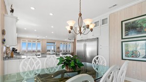 A modern kitchen with white cabinetry, a stainless steel refrigerator, and a dining area with a glass table and white chairs. - Discover Peace on the Beach, a stunning 6BR, 5BA oceanfront retreat in Oak Island. (Oak Island)