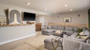 En moderne stue med en grå modulsofa, en træsenggavl, et fladskærms-tv og et spiseområde med et træbord og stole.