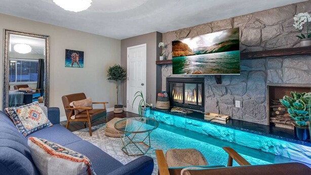 A living room with a stone fireplace, a TV mounted on the wall, a pool with blue lighting, and a comfortable seating area with a sofa and chairs. - Cozy Denver Stay | Grill + Patio | Sloan’s Lake| Firepit (Denver)