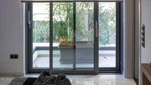 Una habitación moderna con un ventanal corredero que da a un balcón, una cama con manta gris y una mesita de noche de madera.