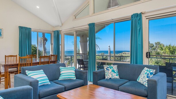 A spacious living area with a large window offering a view of the sea and hills. There are two sofas with patterned cushions and a wooden coffee table. - Breakers 2 2/13 East Street (Crescent Head)