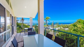 Een balkon met een glazen tafel en stoelen, met uitzicht op een kustlandschap met palmbomen en een zeezicht.