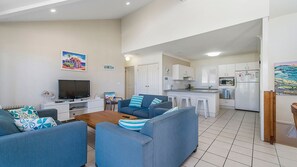 A modern living room with a flat-screen TV, two sofas, a coffee table, and a kitchen area with white cabinets and a microwave.