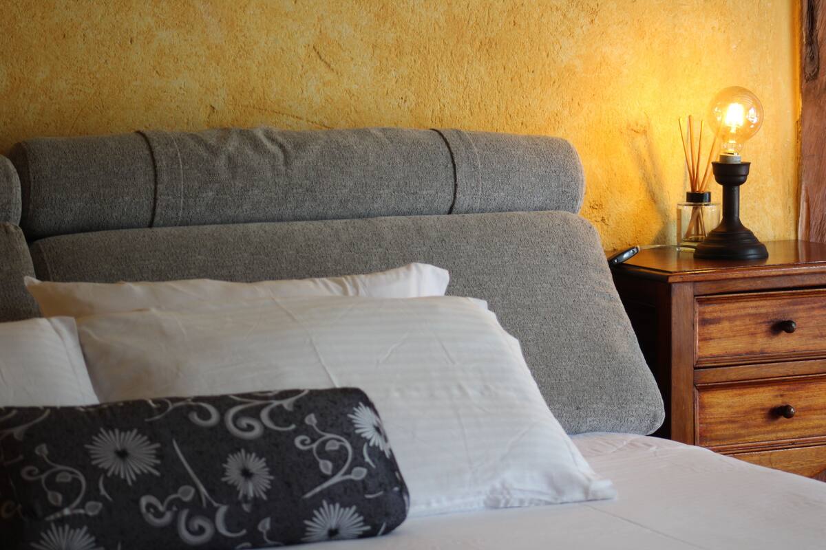 A bed with grey and white bedding, a bedside table with a lamp, and a wooden nightstand.