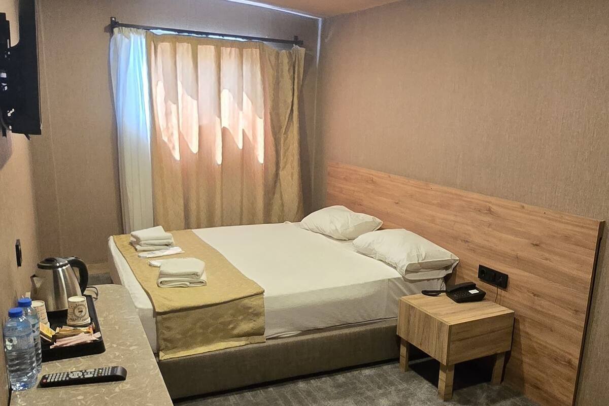 Habitación de hotel con cama, mesita de noche y escritorio con silla. En el escritorio hay controles remotos, una tetera y una botella de agua.