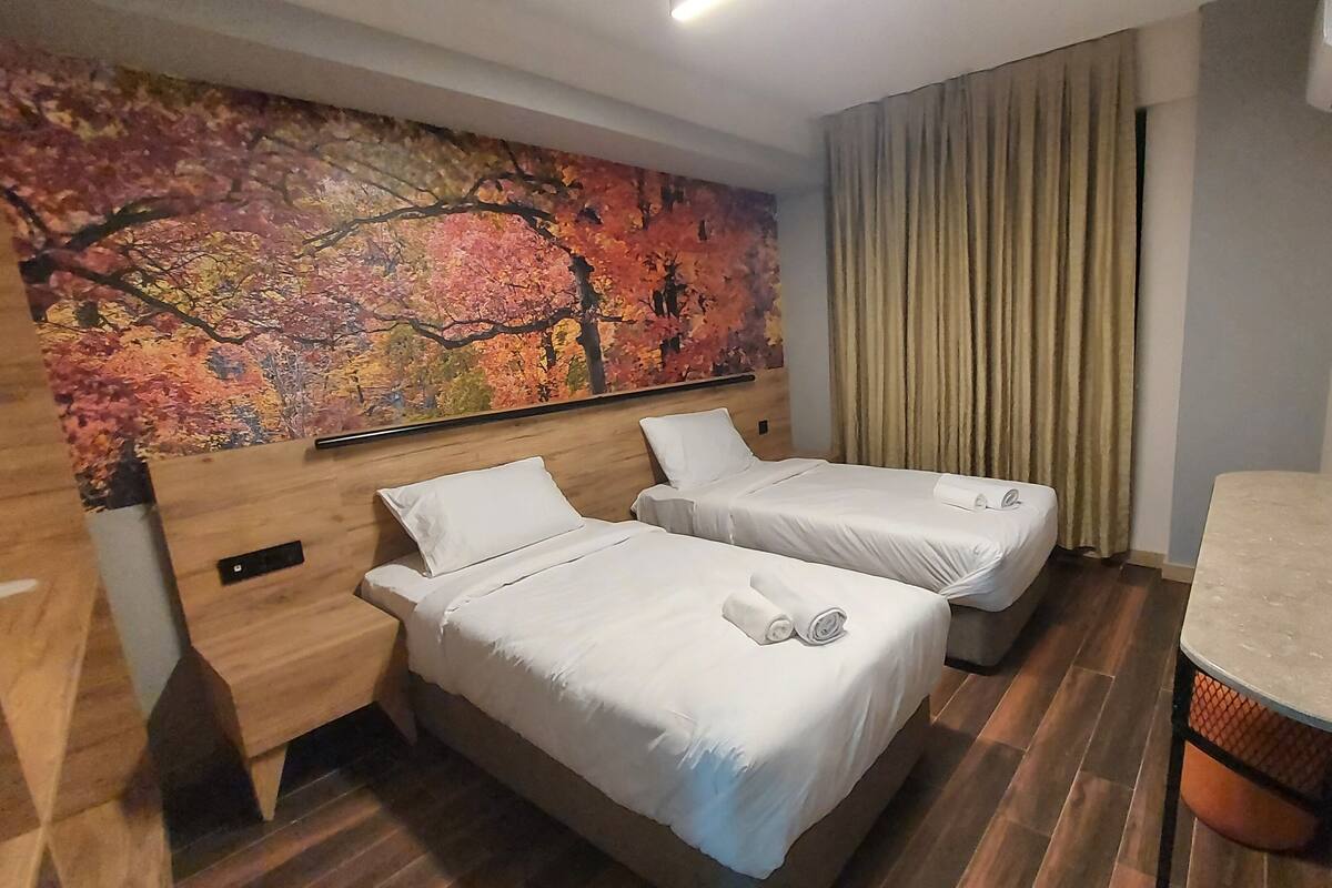 Habitación de hotel con dos camas, un cuadro grande con motivos otoñales en la pared, cabecero de madera, un escritorio pequeño con una silla y un televisor montado en la pared.