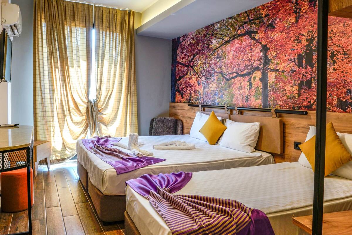Habitación de hotel con dos camas, cabecero de madera, un cuadro grande con motivos otoñales y un escritorio con una silla.