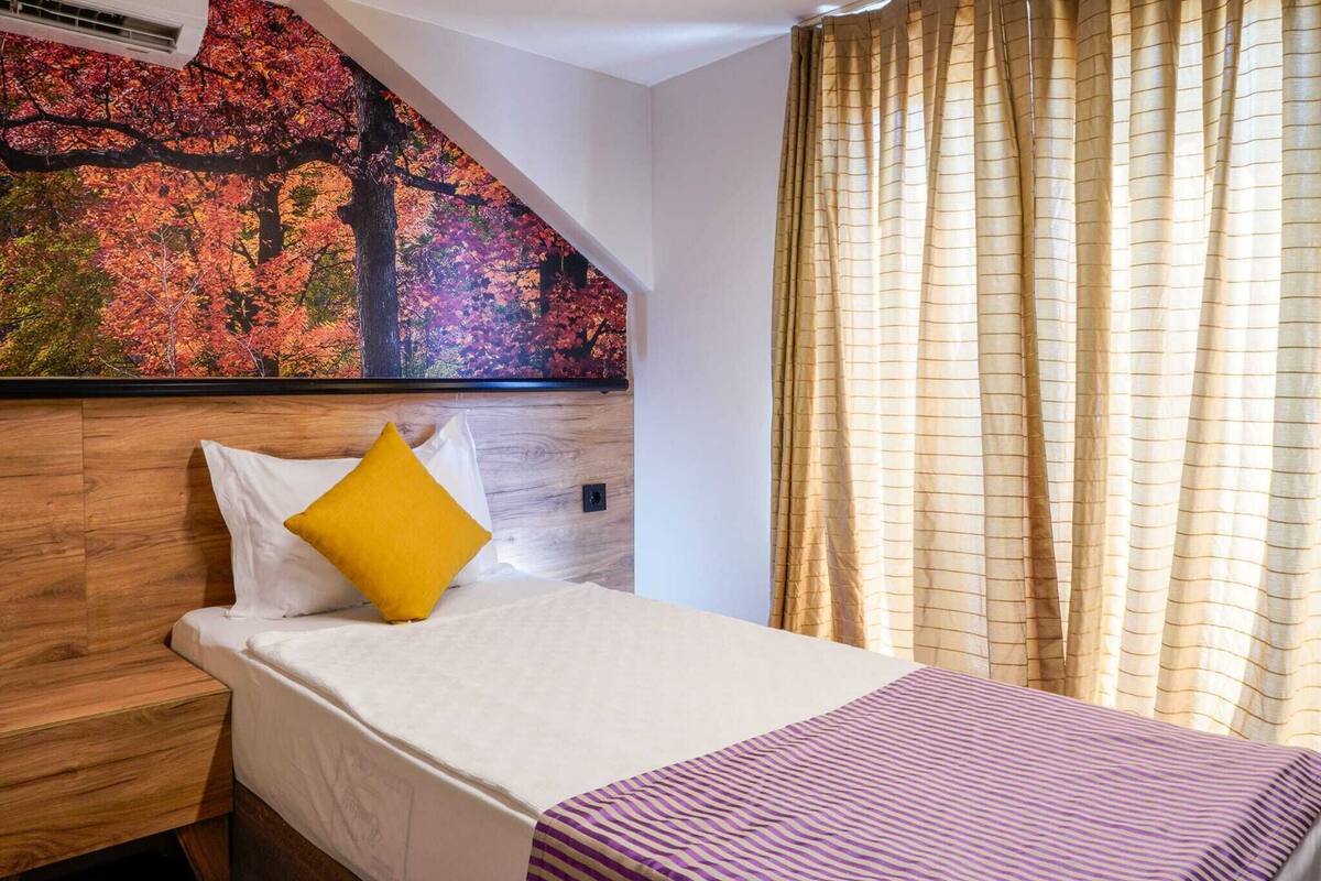 Una habitación de hotel con una cama, cabecero, un cuadro de hojas otoñales y cortinas.