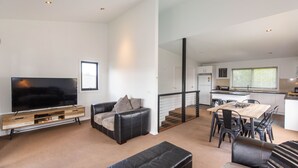 A modern living room with a flat-screen TV, a sofa, and a dining area with a table and chairs. - 25 A Twynam Street, Jindabyne (Jindabyne)