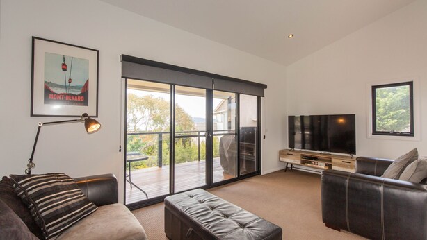 A modern living room with a sofa, ottoman, and a TV on a wooden stand. - 25 A Twynam Street, Jindabyne (Jindabyne)