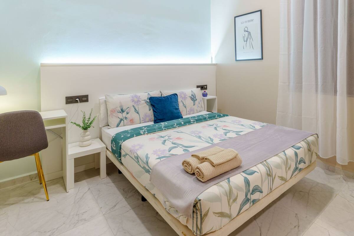 Une chambre à coucher bien rangée, avec un lit, une table de chevet, une chaise et une œuvre d’art accrochée au mur.