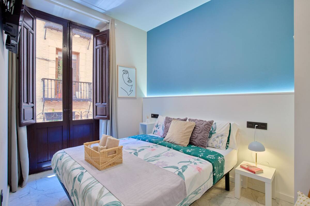 Une chambre à coucher comprenant un lit, une table de chevet, une lampe, une fenêtre avec des rideaux et un mur orné d’un tableau.