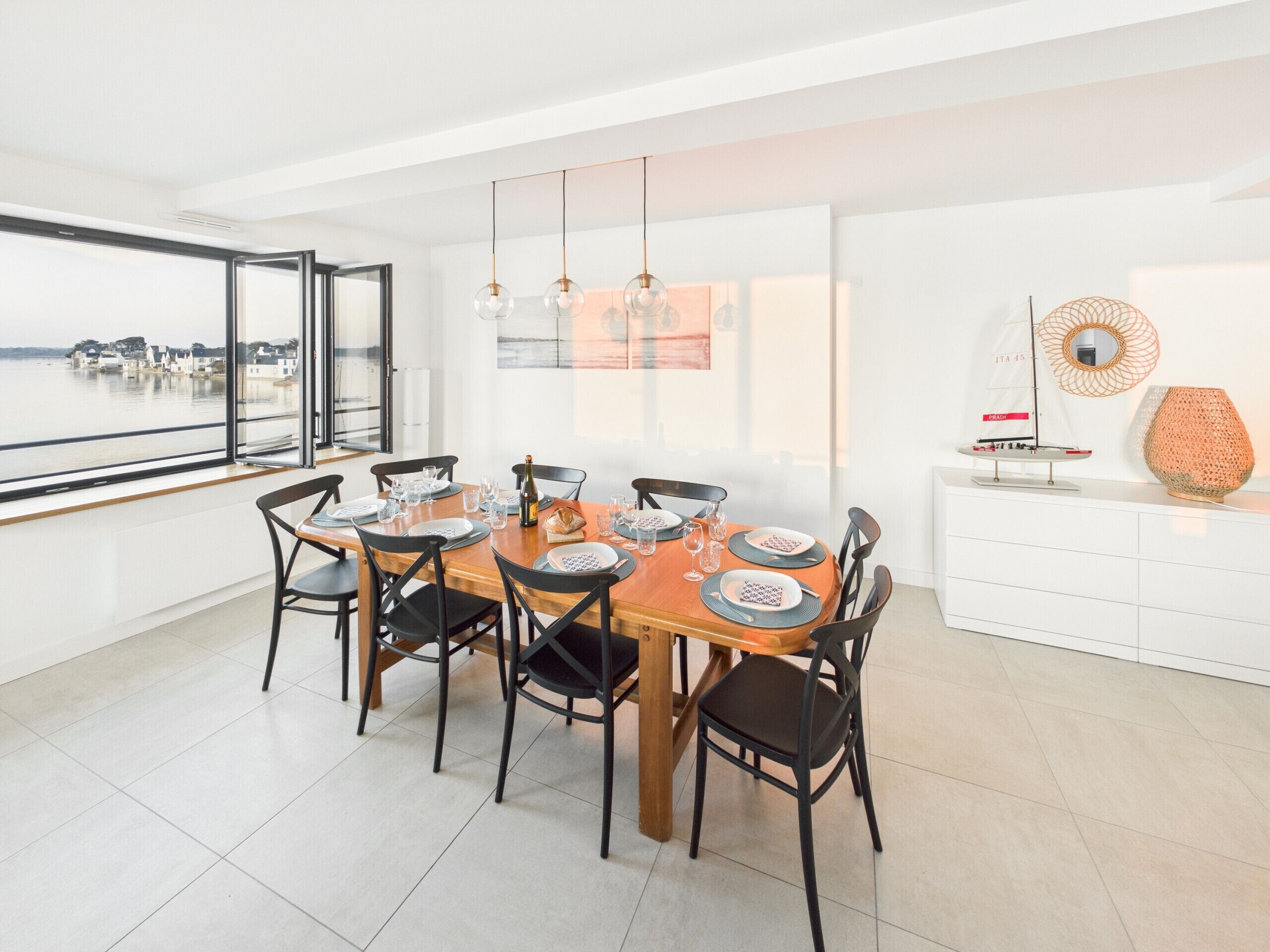 A dining area with a large table set for six, surrounded by chairs, with a view of the sea through large windows.