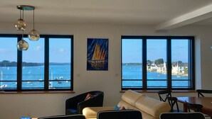 Een woonkamer met grote ramen, een schilderij en uitzicht op zee en gebouwen.