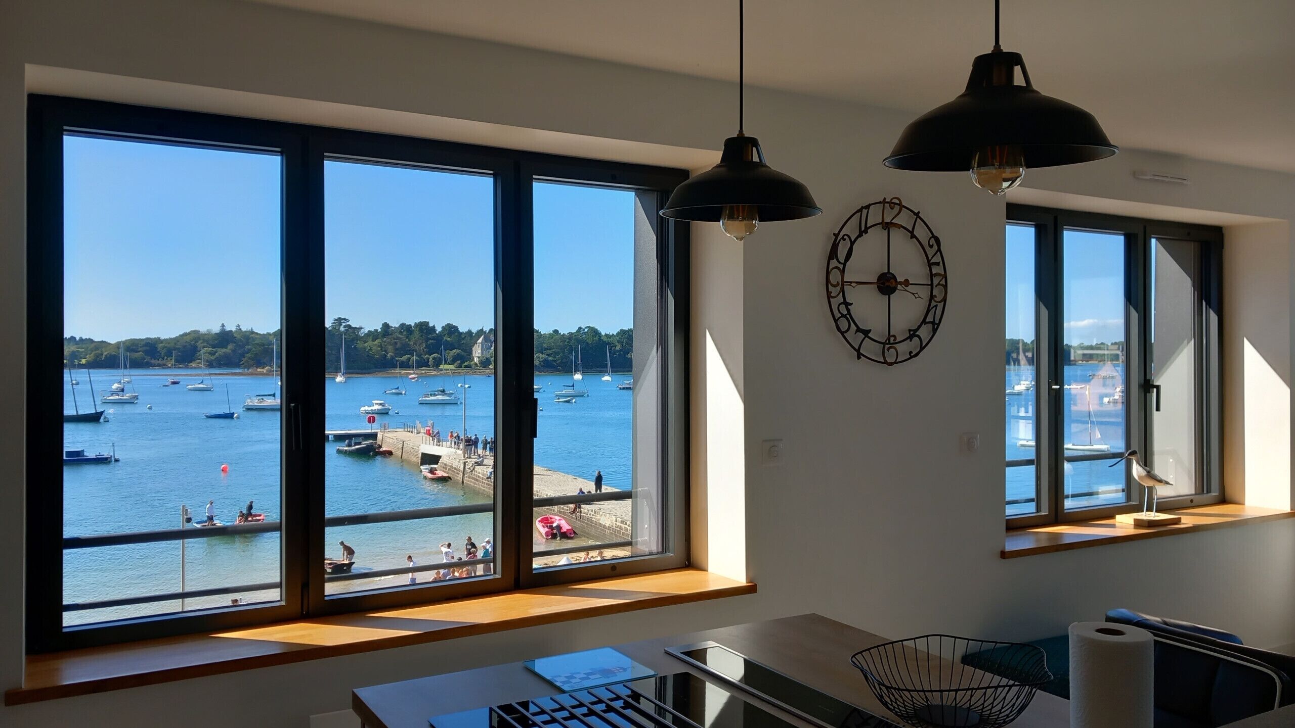 Ein Zimmer mit großen Fenstern mit Blick auf eine Marina, einem Schreibtisch mit einem Laptop und einer Wanduhr.