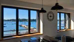 Een kamer met grote ramen met uitzicht op een jachthaven, een bureau met een laptop, en een wandklok.