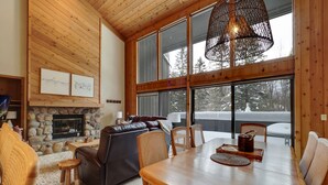 A cozy living room with a stone fireplace, wooden ceiling, and large windows overlooking a snowy landscape. - Hamlet Village Chalet at Nubs Nob/ 4 bedrooms sleeps 12 (Harbor Springs)