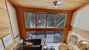 A cozy cabin interior with a wooden ceiling, large windows overlooking a snowy forest, a dining area with wooden furniture, and a fireplace. - Hamlet Village Chalet at Nubs Nob/ 4 bedrooms sleeps 12 (Harbor Springs)