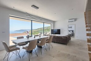 A modern living room with a dining area, large windows, a flat-screen TV, and a staircase. - Villa Ateneum with sea view, jacuzzi&infinity pool (Ripenda Kosi)