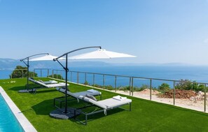 A poolside area with sun loungers and umbrellas, overlooking a body of water and distant mountains.