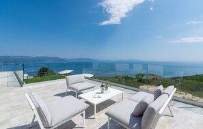 Un espace de détente extérieur moderne, doté d’une balustrade en verre, de mobilier blanc et offrant une vue sur la mer et les collines lointaines.