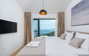 A hotel room with a large bed, a flat-screen TV, a view of the sea, and a painting on the wall.