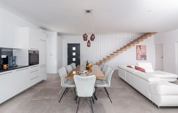 A modern kitchen with white cabinets, a dining table with chairs, a white sofa, and a wooden staircase.
