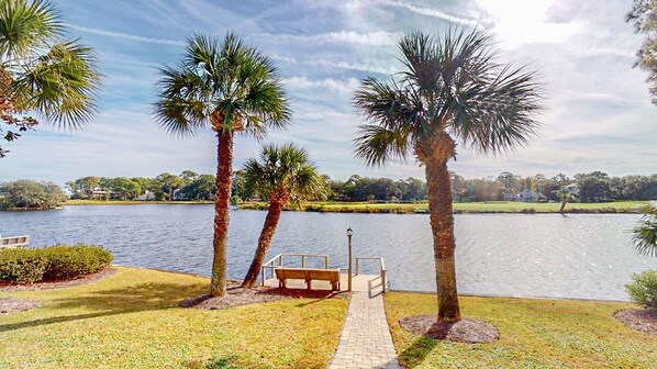 A pathway leading to a bench by a lake with palm trees and a clear sky.