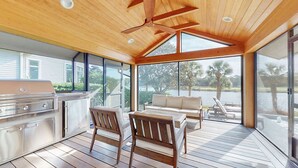 Un amplio espacio de cocina al aire libre con una parrilla, sillas de madera y un sofá, con vistas a una masa de agua.