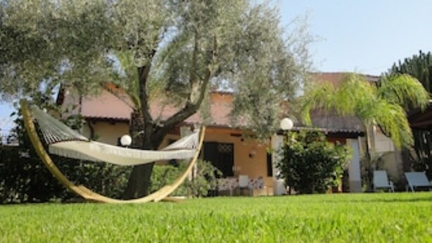 A hammock under a tree in a well-maintained garden with a building in the background. - Villa Paola - Fontane Bianche (Fontane Bianche)