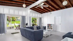 A spacious living area with a large sofa, a coffee table, and a bookshelf. There is a dining area with chairs and a table visible through the glass doors.