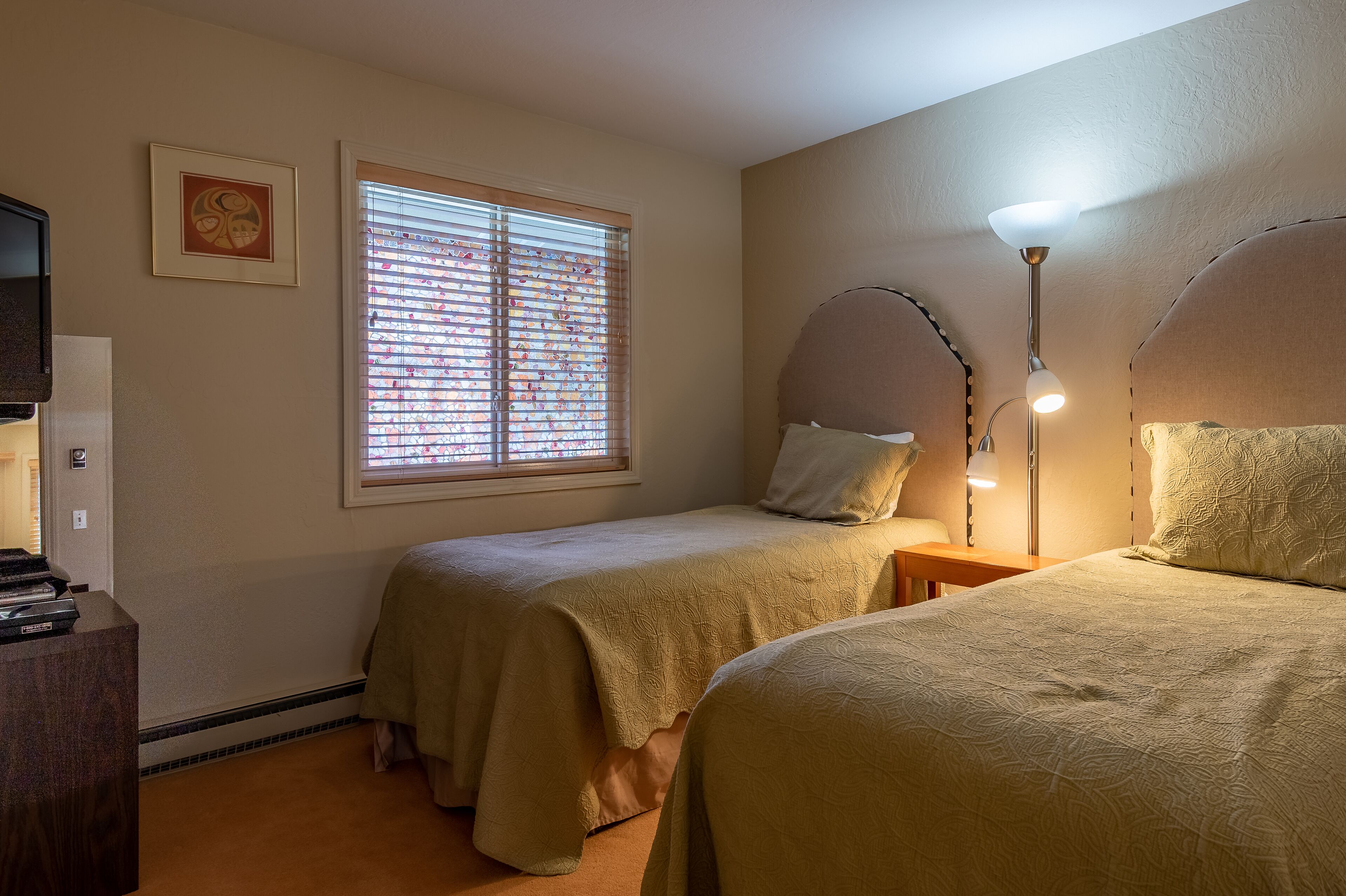 Une chambre avec deux lits, un bureau, une lampe et une fenêtre avec des stores.