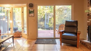 Ein Wohnzimmer mit Kamin, einer Bank, einem Sessel und Blick auf eine Terrasse mit Bäumen.