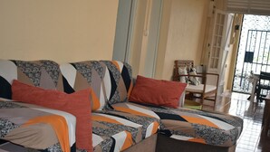 Een bank met een patroon beklede stof en twee rode kussens in een kamer met een betegelde vloer en een deur naar buiten.