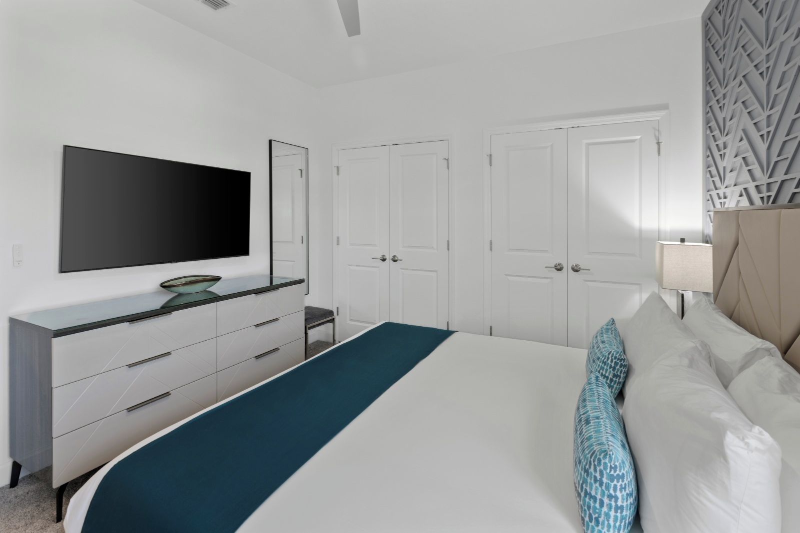 Une chambre à coucher moderne avec un grand lit, un téléviseur fixé au mur, une commode et une porte ornée d’un motif géométrique.