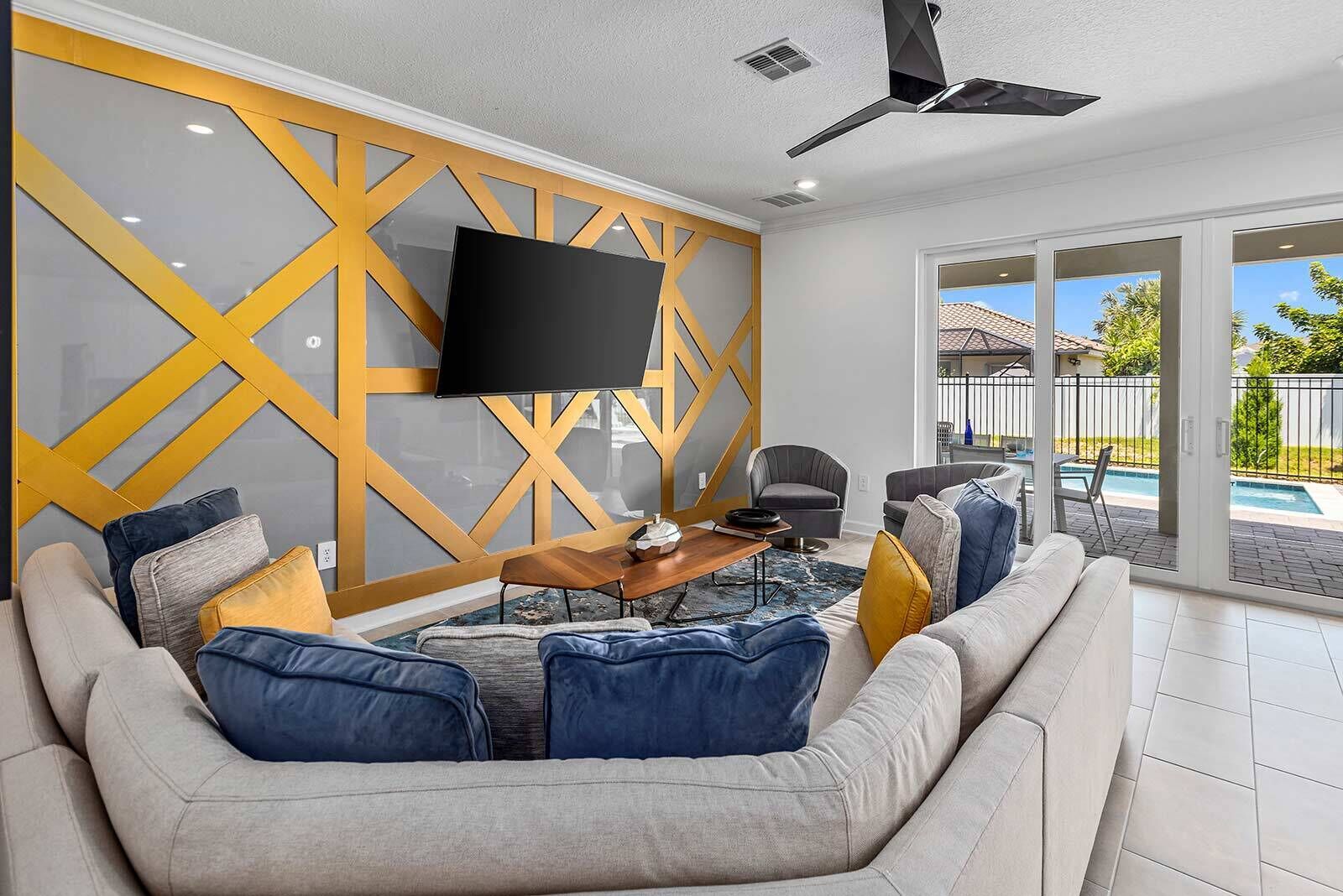 Un salon moderne avec un canapé d’angle, un téléviseur à écran plat fixé sur un mur aux motifs géométriques, un coin repas avec une table et des chaises, et une vue sur une piscine à travers des portes vitrées.