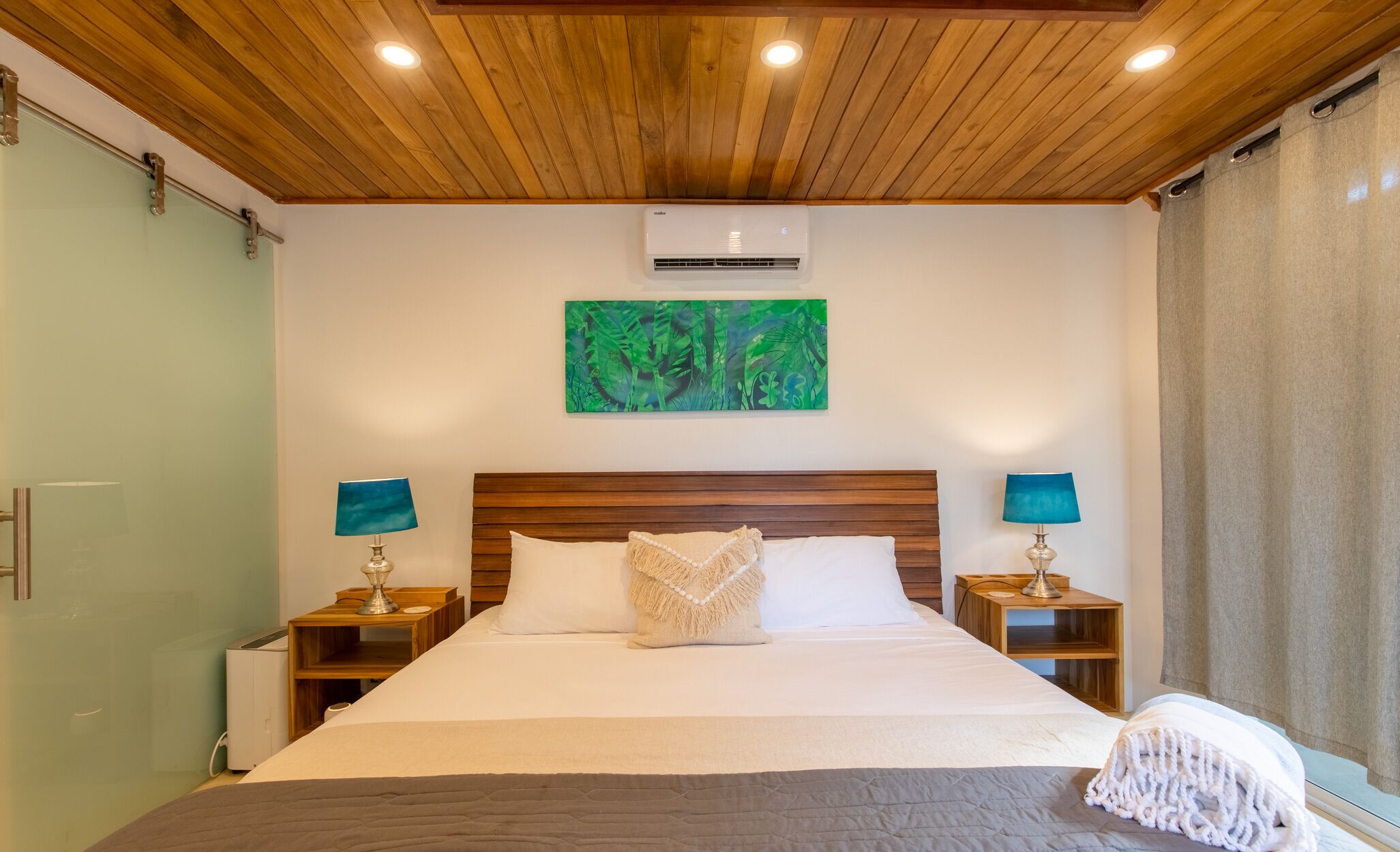 A bedroom with a wooden ceiling, a bed with white bedding, two bedside tables with lamps, and a green abstract painting on the wall.