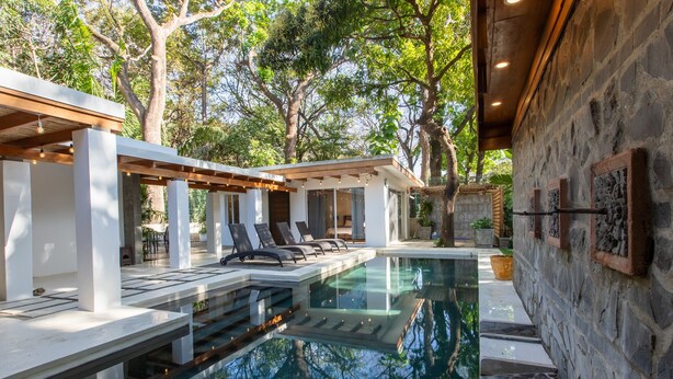 Un área de piscina moderna al aire libre con sillones, una pérgola y un muro de piedra.