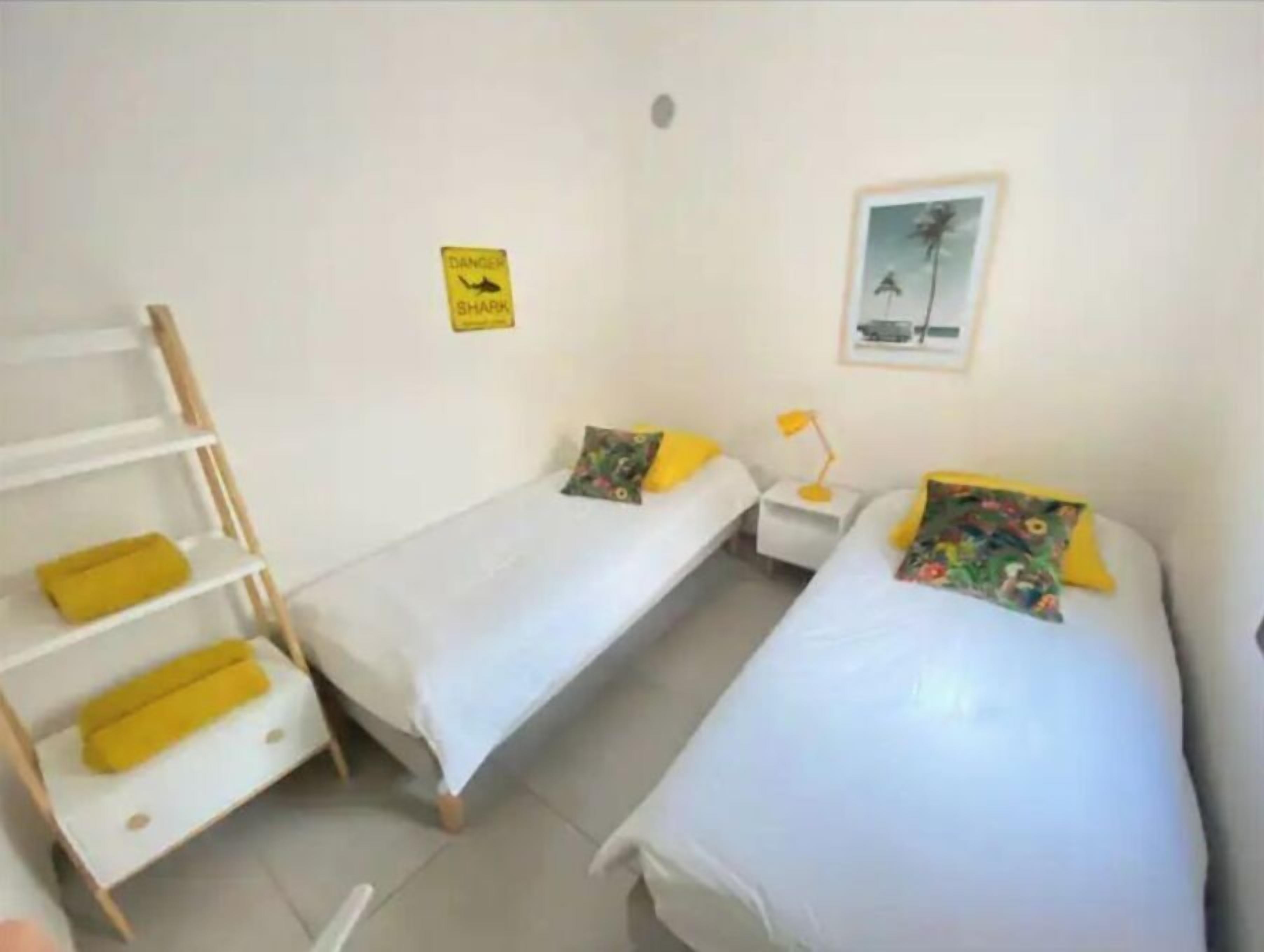 A room with two beds, a shelf with yellow cushions, a small table with a lamp, and a 'Danger Shark' sign on the wall.