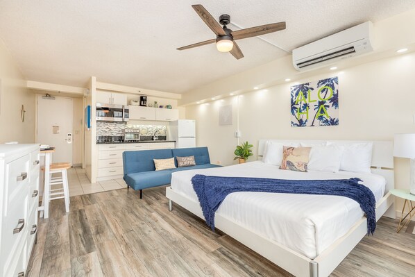 Habitación de hotel compacta con kitchenette, una cama con una manta azul y un ventilador de techo.
