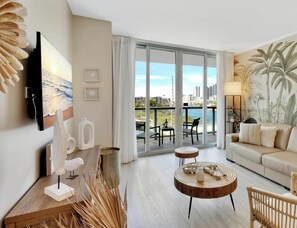 A modern living room with a sofa, a coffee table, a TV mounted on the wall, and a balcony with a view of the city.