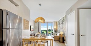 A modern kitchen with a stainless steel refrigerator, a dining table with chairs, a hanging light fixture, and a view of the outdoors through large windows.