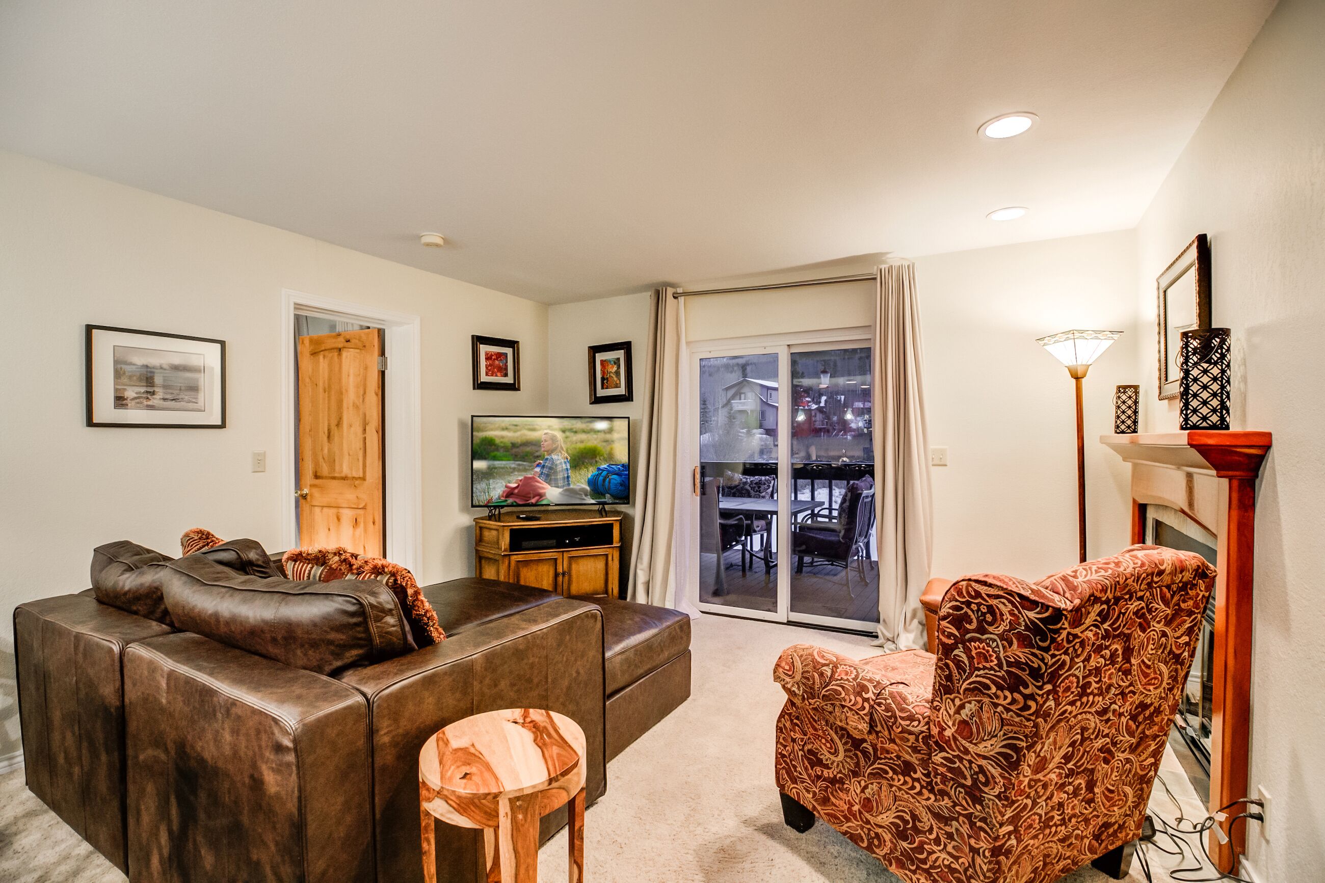 A living room with a brown leather sofa, a patterned armchair, a television, and a sliding glass door leading to an outdoor patio.